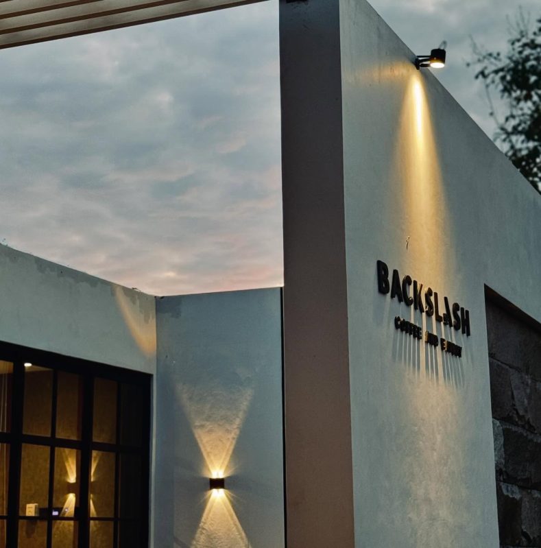 Dinding luar BackSlash Coffee and Eatery saat senja dengan lampu dinding menyala